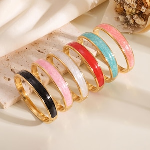 Op de afbeelding: Een set van zes scharnierende armbanden met goudkleurige metalen randen en gekleurde emaille centra. De armbanden zijn verkrijgbaar in zwart, lichtroze, wit, rood, turquoise en zachtroze, elk met een subtiel wervelend patroon.