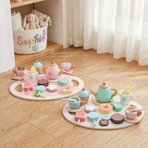 May include: Wooden toy tea sets with pink and mint green teapots, cups, and treats on oval trays. The sets include miniature cakes, ice cream, and cookies. A basket with the name "Sophie" is in the background.