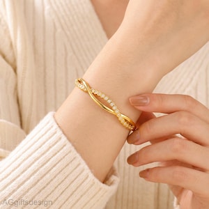 May include: A gold-toned bangle bracelet featuring a dual-strand design. One strand is a smooth, polished gold-colored metal, while the other is a delicate chain set with a series of small, clear, round-cut stones that follow a gentle wave pattern.