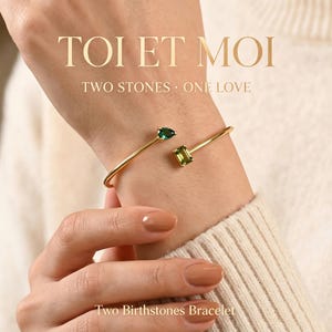 Pode incluir: Uma pulseira aberta de tom dourado com duas pedras preciosas distintas: uma pedra verde-escura em corte de pera e uma pedra verde-clara retangular em corte esmeralda. O texto TOI ET MOI TWO STONES ONE LOVE e Two Birthstones Bracelet é visível.