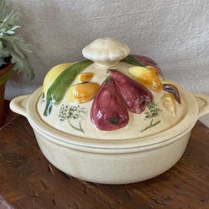May include: Cream-colored ceramic casserole dish with a lid featuring colorful, raised vegetable designs. The lid has a mushroom-shaped handle. The dish sits on a wooden surface, suggesting a rustic kitchen theme.