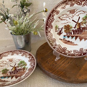 May include: Two antique ceramic plates with a brown and white windmill scene. The plates have a decorative brown border. A small metal bucket with white flowers is in the background. The plates are displayed on a wooden surface.