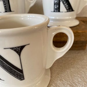 May include: White ceramic mugs with black letter designs. The mugs have a classic shape with a handle and a wide base. The letter "N" is prominently displayed on the front of one mug. Other mugs with letters are visible in the background.