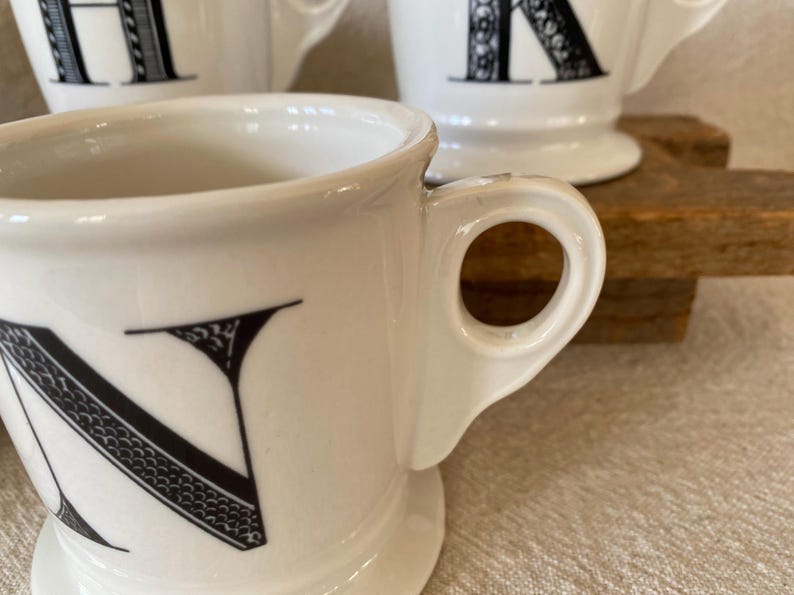 May include: White ceramic mugs with black letter designs. The mugs have a classic shape with a handle and a wide base. The letter "N" is prominently displayed on the front of one mug. Other mugs with letters are visible in the background.