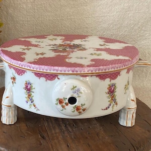 May include: A white and pink porcelain serving dish with a lid. The lid and body are decorated with floral patterns. The dish has a round shape with a handle on each side and is supported by three small legs.