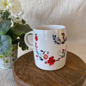 Puede incluir: Taza de cer&aacute;mica blanca con asa curva, adornada con un dise&ntilde;o floral en rojo, azul y verde. La taza est&aacute; sobre una superficie de madera, con una planta en maceta en el fondo.