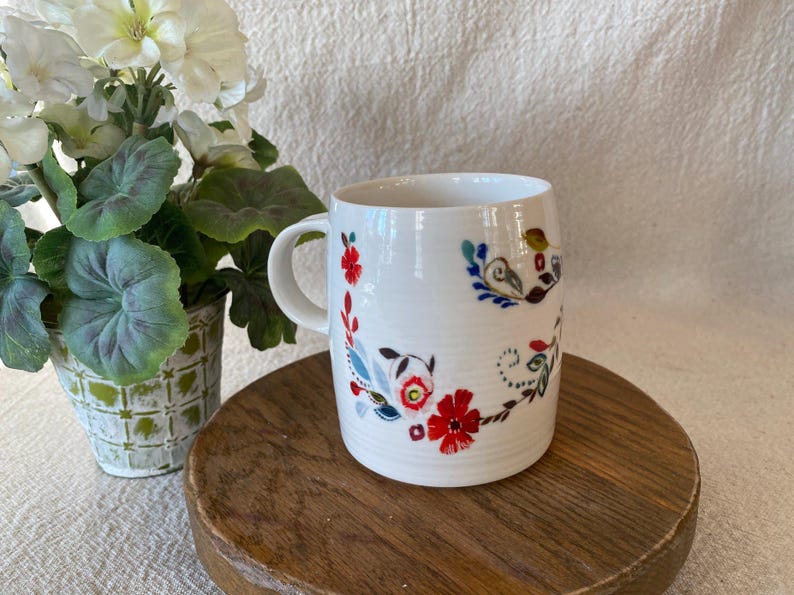 Puede incluir: Taza de cer&aacute;mica blanca con asa curva, adornada con un dise&ntilde;o floral en rojo, azul y verde. La taza est&aacute; sobre una superficie de madera, con una planta en maceta en el fondo.
