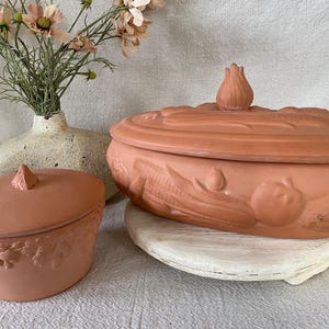 May include: Two terracotta-colored ceramic serving dishes with lids. The larger dish has a tulip-shaped handle and embossed vegetables. The smaller dish has a triangular handle and embossed fruit. A vase with flowers is in the background.