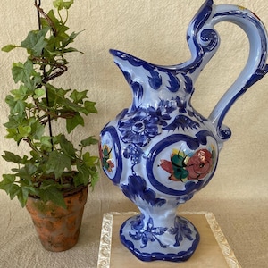 May include: A hand-painted ceramic pitcher with a blue and white floral design. The pitcher features a curved handle and a spout. The design includes red, green, and yellow floral accents. A small potted plant with green leaves is to the left of the pitcher.