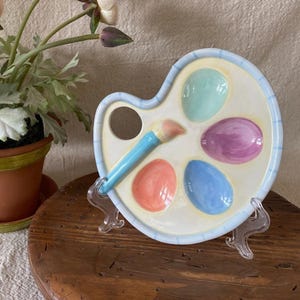 May include: A ceramic artist palette dish featuring a cream-colored base with a light blue striped border. The palette includes four recessed oval wells painted in pastel mint green, lavender, peach, and sky blue, with a sculpted paintbrush handle.