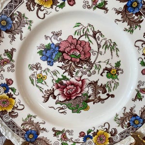 May include: A white ceramic plate with a floral design. The plate features a central arrangement of pink, blue, and yellow flowers with brown and green foliage. The rim has a brown border and a white, scalloped edge. A decorative piece for the dining table.
