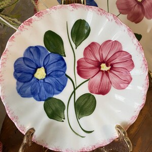 May include: A white ceramic plate with a scalloped edge featuring a hand-painted design of one blue flower and one pink flower with green leaves. The rim of the plate is decorated with a thin, textured pink border that follows the scalloped shape.