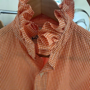 Peut inclure: Une chemise boutonn&eacute;e &agrave; motif vichy orange et blanc avec un col haut &agrave; volants. La chemise pr&eacute;sente une patte de boutonnage sur le devant et un petit logo brod&eacute; vert sur la poitrine. Le tissu arbore un motif &agrave; carreaux classique.