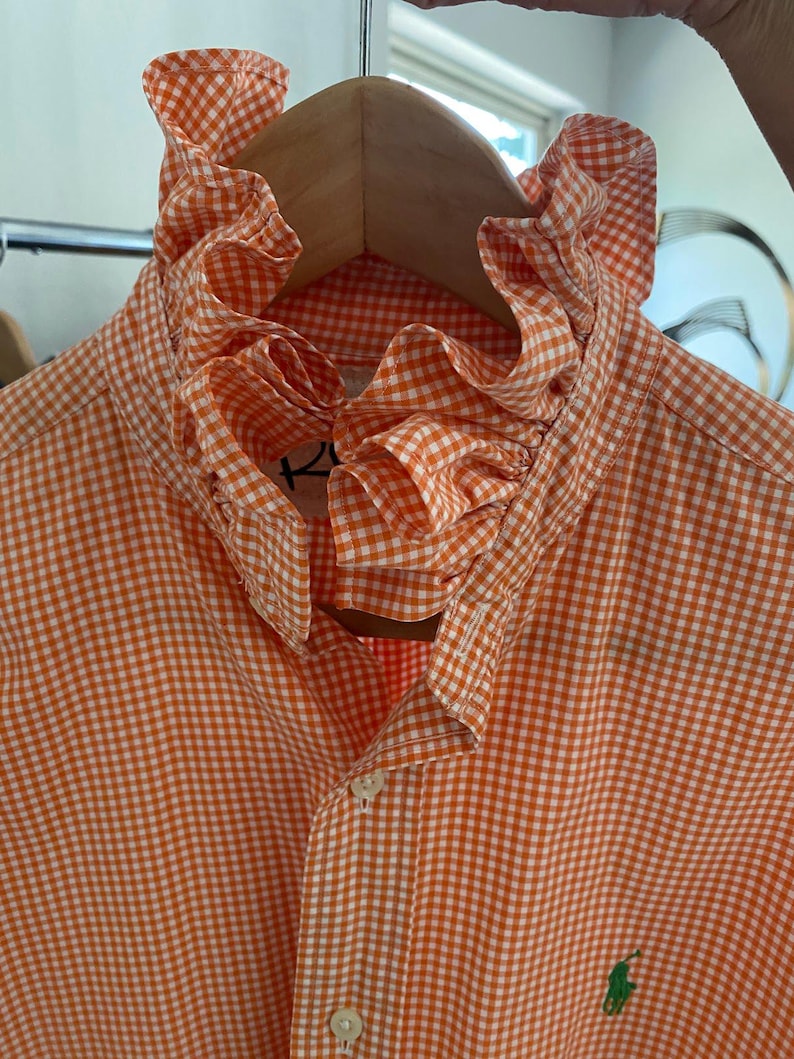 Peut inclure: Une chemise boutonn&eacute;e &agrave; motif vichy orange et blanc avec un col haut &agrave; volants. La chemise pr&eacute;sente une patte de boutonnage sur le devant et un petit logo brod&eacute; vert sur la poitrine. Le tissu arbore un motif &agrave; carreaux classique.