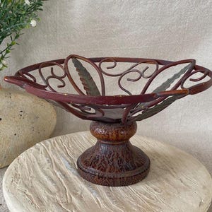 May include: A decorative, ornate metal bowl with a pedestal base. The bowl is a deep reddish-brown color with a leaf and scroll design. The pedestal is a darker brown with a textured finish. The bowl is likely used for decorative purposes.