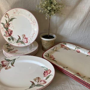 May include: A collection of white ceramic dinnerware with a floral tulip design. The set includes plates and a rectangular serving tray, all with a red trim. A small potted plant is in the background.