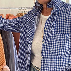 Peut inclure: Une chemise boutonn&eacute;e &agrave; carreaux vichy bleus et blancs avec un col &agrave; volants, des manches longues et une poche poitrine. La chemise est port&eacute;e sur un t-shirt blanc uni et un jean bleu clair, avec un fin collier dor&eacute; visible au niveau du cou.
