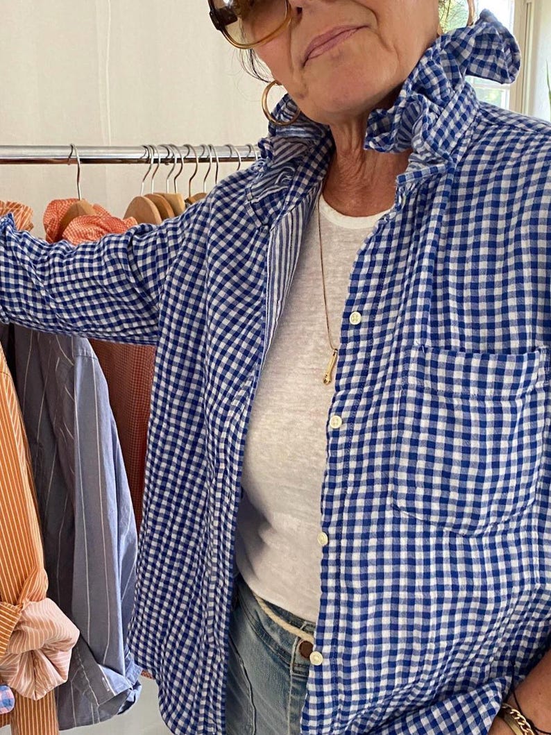 Peut inclure: Une chemise boutonn&eacute;e &agrave; carreaux vichy bleus et blancs avec un col &agrave; volants, des manches longues et une poche poitrine. La chemise est port&eacute;e sur un t-shirt blanc uni et un jean bleu clair, avec un fin collier dor&eacute; visible au niveau du cou.