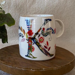 Puede incluir: Taza de cer&aacute;mica blanca con un gran dise&ntilde;o floral "K" colorido. La letra est&aacute; adornada con patrones florales rojos, azules y verdes. La taza tiene un asa curva y est&aacute; colocada sobre una superficie de madera.