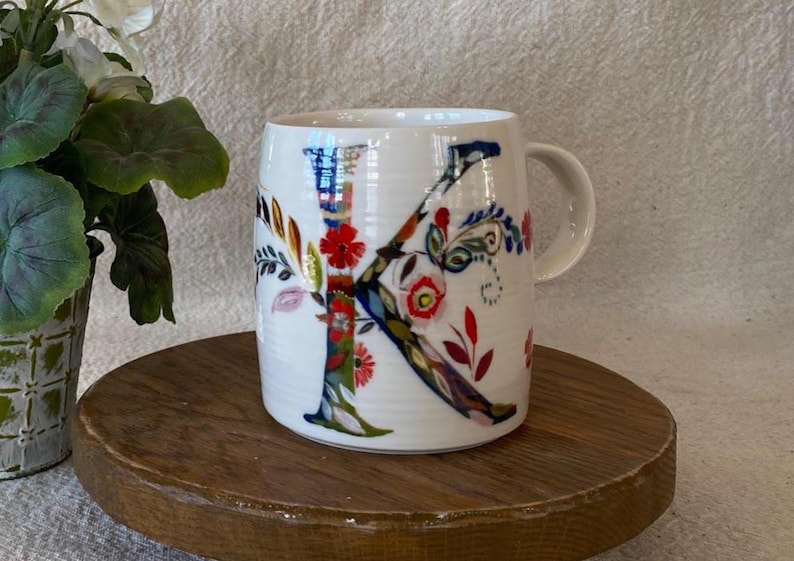 Puede incluir: Taza de cer&aacute;mica blanca con un gran dise&ntilde;o floral "K" colorido. La letra est&aacute; adornada con patrones florales rojos, azules y verdes. La taza tiene un asa curva y est&aacute; colocada sobre una superficie de madera.