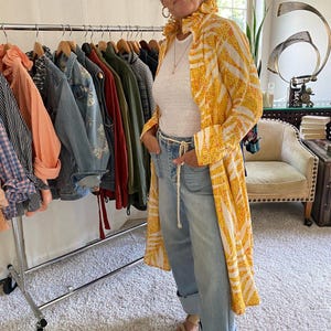 Peut inclure: Un long manteau jaune et blanc &agrave; motifs avec un col &agrave; volants, port&eacute; sur un t-shirt blanc et un jean bleu clair avec une ceinture en corde. Le manteau pr&eacute;sente un imprim&eacute; r&eacute;p&eacute;titif en forme de feuilles et est port&eacute; ouvert sur la tenue.