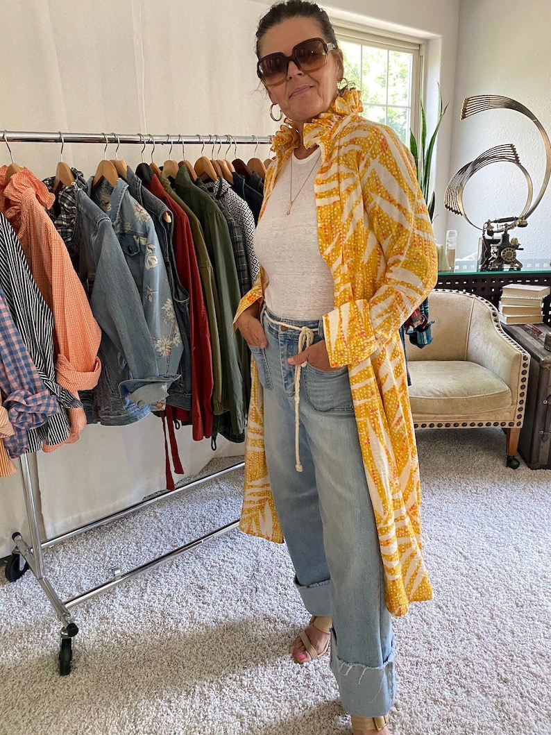 Peut inclure: Un long manteau jaune et blanc &agrave; motifs avec un col &agrave; volants, port&eacute; sur un t-shirt blanc et un jean bleu clair avec une ceinture en corde. Le manteau pr&eacute;sente un imprim&eacute; r&eacute;p&eacute;titif en forme de feuilles et est port&eacute; ouvert sur la tenue.