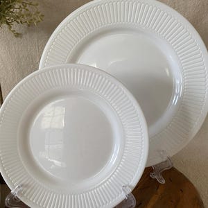 May include: Two white ceramic plates of different sizes. Both plates have a ridged border design. The plates are displayed on clear plastic stands. The plates are on a wooden surface.