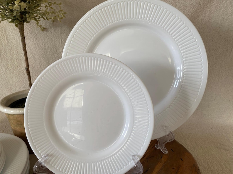 May include: Two white ceramic plates of different sizes. Both plates have a ridged border design. The plates are displayed on clear plastic stands. The plates are on a wooden surface.