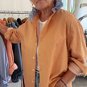 Peut inclure: Une chemise boutonn&eacute;e &agrave; rayures verticales orange et blanches avec un d&eacute;tail de col &agrave; volants ray&eacute; bleu et blanc contrastant, des manches longues &agrave; poignets boutonn&eacute;s, port&eacute;e sur un sous-v&ecirc;tement blanc avec un jean en denim bleu clair.