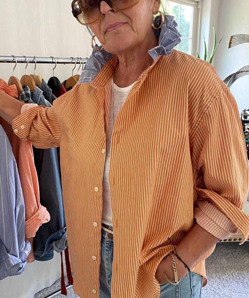 Peut inclure: Une chemise boutonn&eacute;e &agrave; rayures verticales orange et blanches avec un d&eacute;tail de col &agrave; volants ray&eacute; bleu et blanc contrastant, des manches longues &agrave; poignets boutonn&eacute;s, port&eacute;e sur un sous-v&ecirc;tement blanc avec un jean en denim bleu clair.