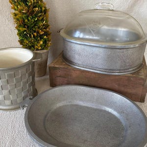 May include: A collection of vintage metal kitchenware, including a casserole dish with a glass lid, an oval serving dish, and a woven-patterned ice bucket. The items are silver-toned and appear to be made of metal.