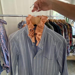 Peut inclure: Une chemise &agrave; manches longues boutonn&eacute;e bleu avec des rayures verticales blanches, dot&eacute;e d'un col &agrave; volants orange et blanc ray&eacute;. La chemise comprend une poche poitrine unique et une fermeture boutonn&eacute;e classique sur le devant.