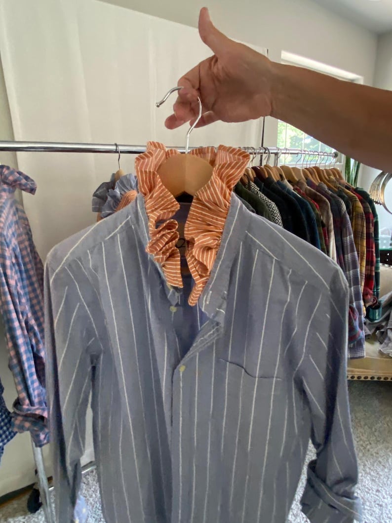 Peut inclure: Une chemise &agrave; manches longues boutonn&eacute;e bleu avec des rayures verticales blanches, dot&eacute;e d'un col &agrave; volants orange et blanc ray&eacute;. La chemise comprend une poche poitrine unique et une fermeture boutonn&eacute;e classique sur le devant.
