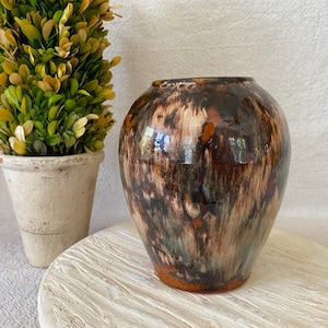 May include: A decorative, glazed ceramic vase with a rounded shape. The vase features a blend of brown, orange, and green hues, creating a marbled effect. It sits on a white, textured surface, with a small potted plant in the background.