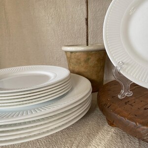 May include: A stack of white ceramic plates with a ribbed design around the edges. The plates are arranged on a wooden surface, with a few plates displayed on a clear stand. A small, decorative pot is in the background.