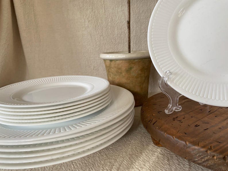 May include: A stack of white ceramic plates with a ribbed design around the edges. The plates are arranged on a wooden surface, with a few plates displayed on a clear stand. A small, decorative pot is in the background.