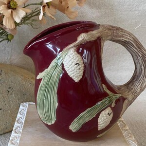 May include: A deep burgundy ceramic pitcher featuring a textured handle shaped like a tree branch. The body is decorated with raised, white pinecones and green pine needles. The pitcher has a glossy finish and a wide, curved pouring spout.