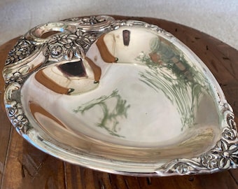 May include: A silver-colored, heart-shaped dish with ornate floral and scrollwork detailing around the rim and handle. The dish has a reflective surface, and is likely made of metal. The dish is sitting on a wooden surface.