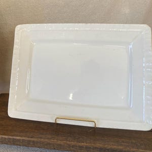 May include: A rectangular, white ceramic serving dish with a slightly raised, textured border. The dish is supported by a gold-colored metal stand, and sits on a dark wooden surface. The overall aesthetic is clean and classic.