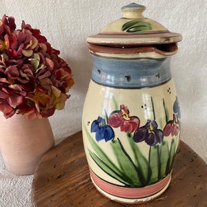 May include: A hand-painted ceramic pitcher with a lid, featuring iris flower designs in blue, red, and purple. The pitcher has a cream-colored base, blue band, and a pink and brown rim. A vase with flowers is to the left.