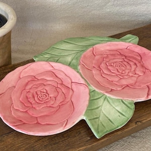 May include: Two pink rose-shaped ceramic dishes rest on a green leaf-shaped base. The dishes are a light pink color with detailed rose patterns. The base is green, and the items are on a wooden surface.