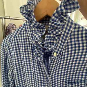 Peut inclure: Une chemise &agrave; manches longues boutonn&eacute;e avec un motif &agrave; carreaux vichy bleu et blanc. La chemise pr&eacute;sente un col &agrave; volants pro&eacute;minent et une poche poitrine unique. Elle est faite d'un tissu l&eacute;ger et comporte de petits boutons blancs sur le devant.