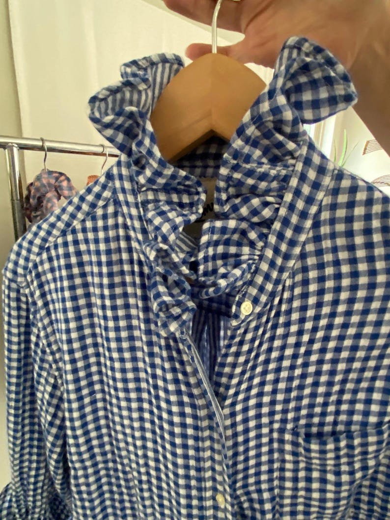 Peut inclure: Une chemise &agrave; manches longues boutonn&eacute;e avec un motif &agrave; carreaux vichy bleu et blanc. La chemise pr&eacute;sente un col &agrave; volants pro&eacute;minent et une poche poitrine unique. Elle est faite d'un tissu l&eacute;ger et comporte de petits boutons blancs sur le devant.