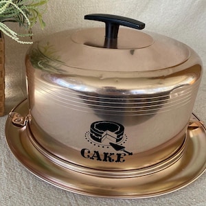 May include: A copper-toned cake carrier with a black handle and a matching base. The carrier features a graphic of a cake with a slice cut out and the word "CAKE" below it. The carrier is designed to hold and transport cakes.