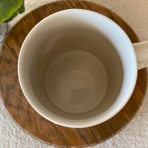Puede incluir: Una taza de cer&aacute;mica blanca con asa, colocada sobre un posavasos redondo de madera. El interior de la taza es de color beige claro. El posavasos tiene un patr&oacute;n de vetas de madera natural. Una peque&ntilde;a planta verde es visible en el fondo.