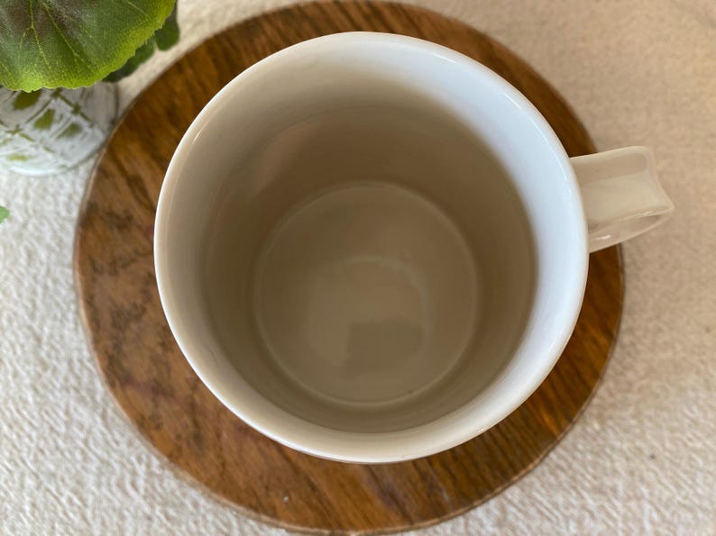 Puede incluir: Una taza de cer&aacute;mica blanca con asa, colocada sobre un posavasos redondo de madera. El interior de la taza es de color beige claro. El posavasos tiene un patr&oacute;n de vetas de madera natural. Una peque&ntilde;a planta verde es visible en el fondo.