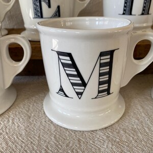 May include: White ceramic mug featuring the letter "M" in black, with a striped pattern. The mug has a rounded handle and a wide base. Other similar mugs are visible, indicating a set. The mugs are made of ceramic.