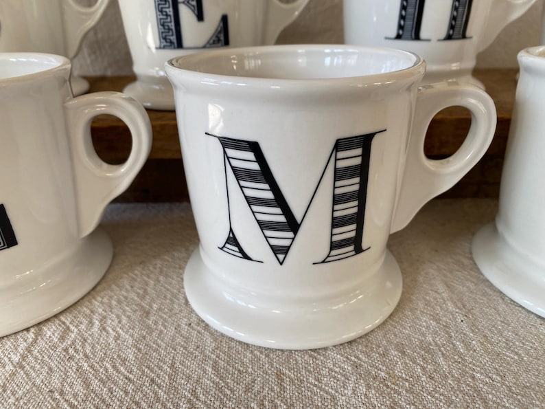 May include: White ceramic mug featuring the letter "M" in black, with a striped pattern. The mug has a rounded handle and a wide base. Other similar mugs are visible, indicating a set. The mugs are made of ceramic.