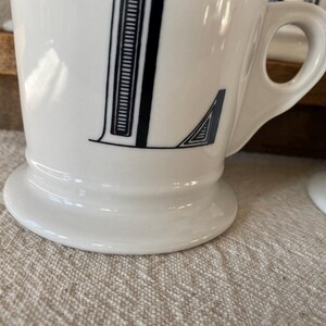 May include: White ceramic mug featuring a black letter "L". The letter is designed with a vertical stripe pattern and a geometric detail. The mug has a rounded base and a handle.