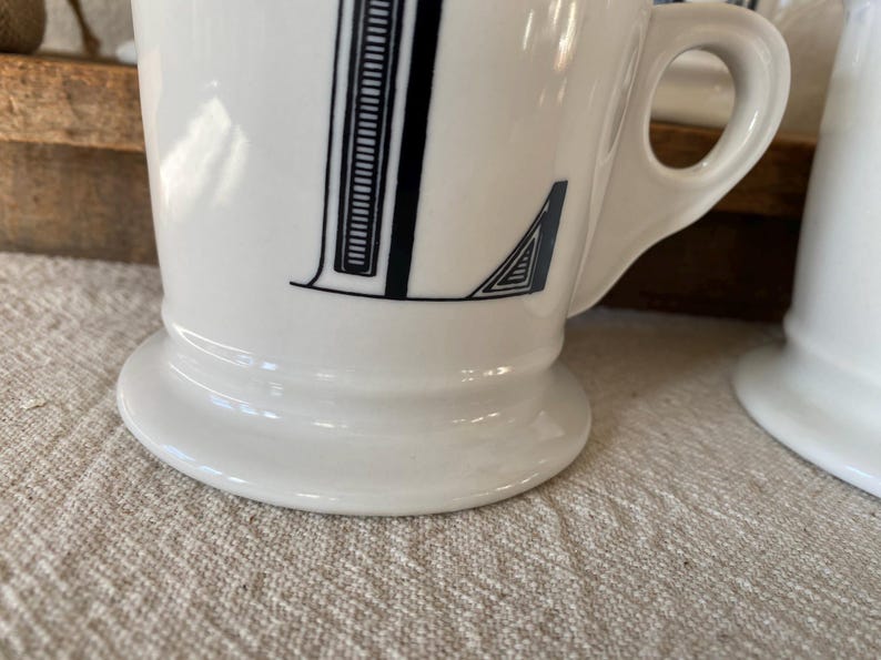 May include: White ceramic mug featuring a black letter "L". The letter is designed with a vertical stripe pattern and a geometric detail. The mug has a rounded base and a handle.